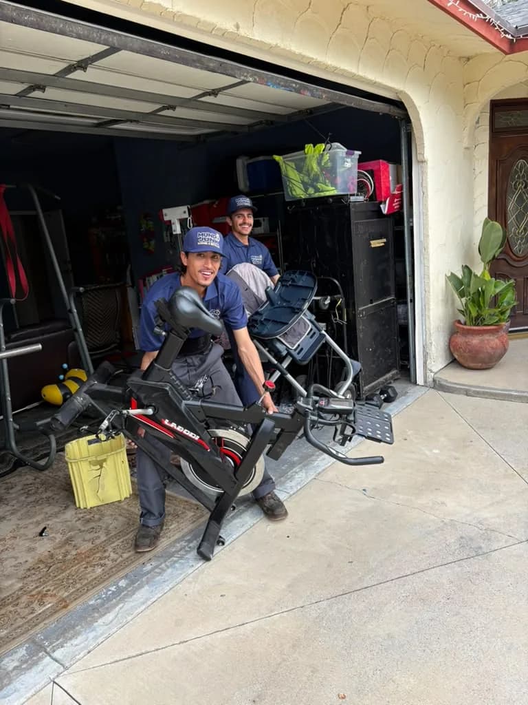 Mundo Junk Removal & Recycling crew carrying an exercise bike and gym equipment out of a garage